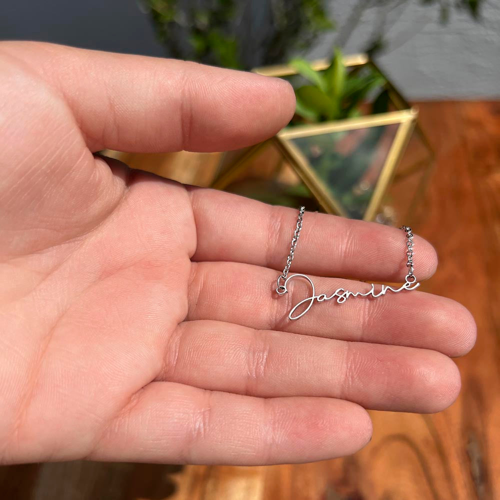 Hand holding the Soft Life Society Signature Style Name Necklace in silver with “Jasmine” script pendant, shown in natural light with a geometric glass planter in the background.