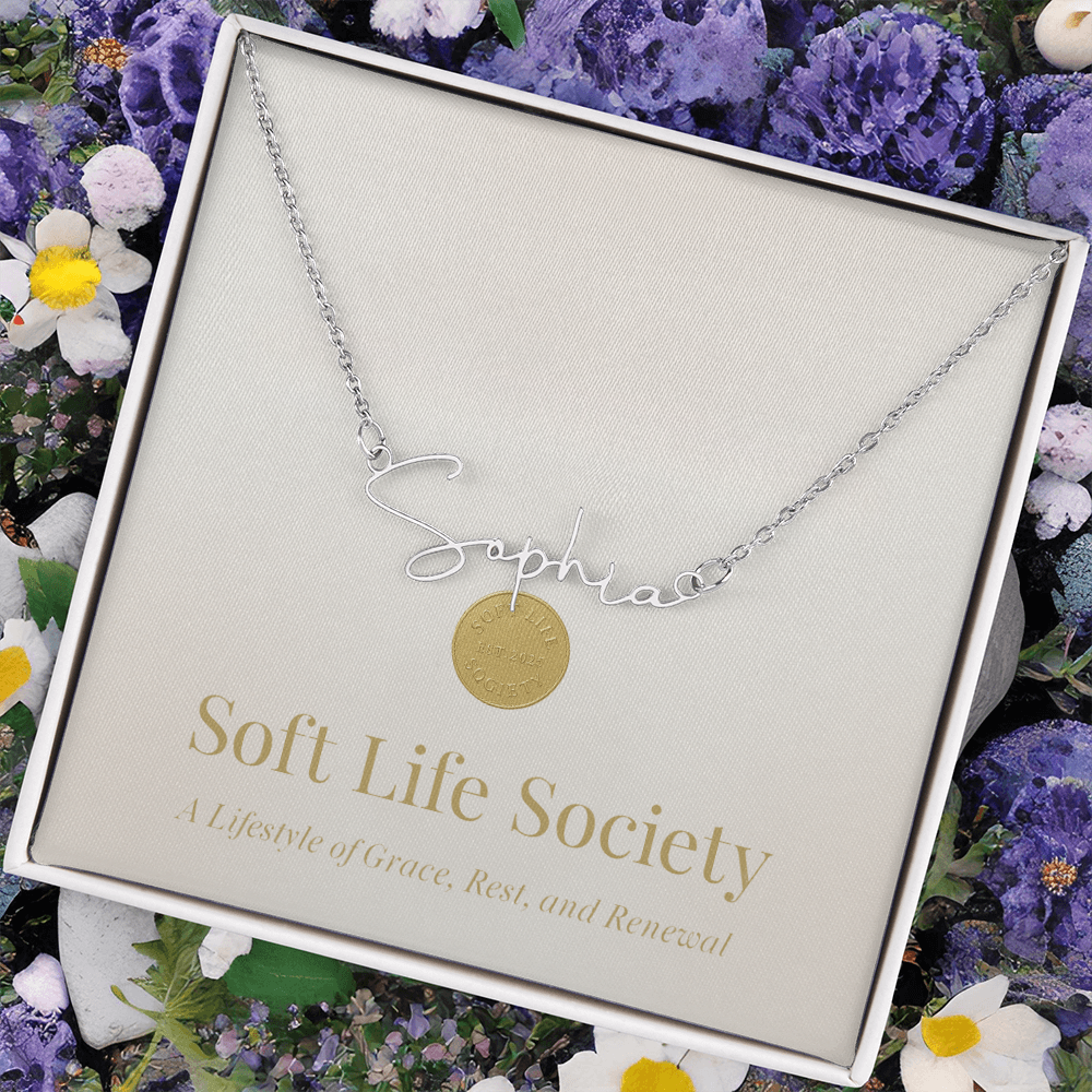 Soft Life Society Signature Style Name Necklace in silver with “Sophia” script pendant, displayed in a gift box surrounded by purple and white flowers for a soft, elegant presentation.