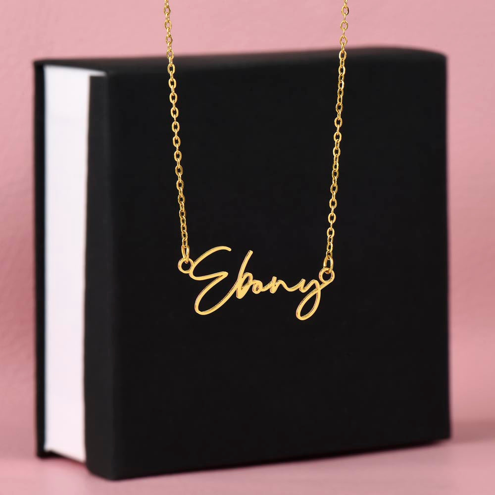 Soft Life Society Signature Style Name Necklace in 18K yellow gold with “Ebony” script pendant displayed in front of a black standard gift box on a blush pink background.