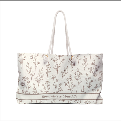 Floral weekender travel bag with taupe botanical pattern held by woman in elegant cream dress in sunlit neutral room.
