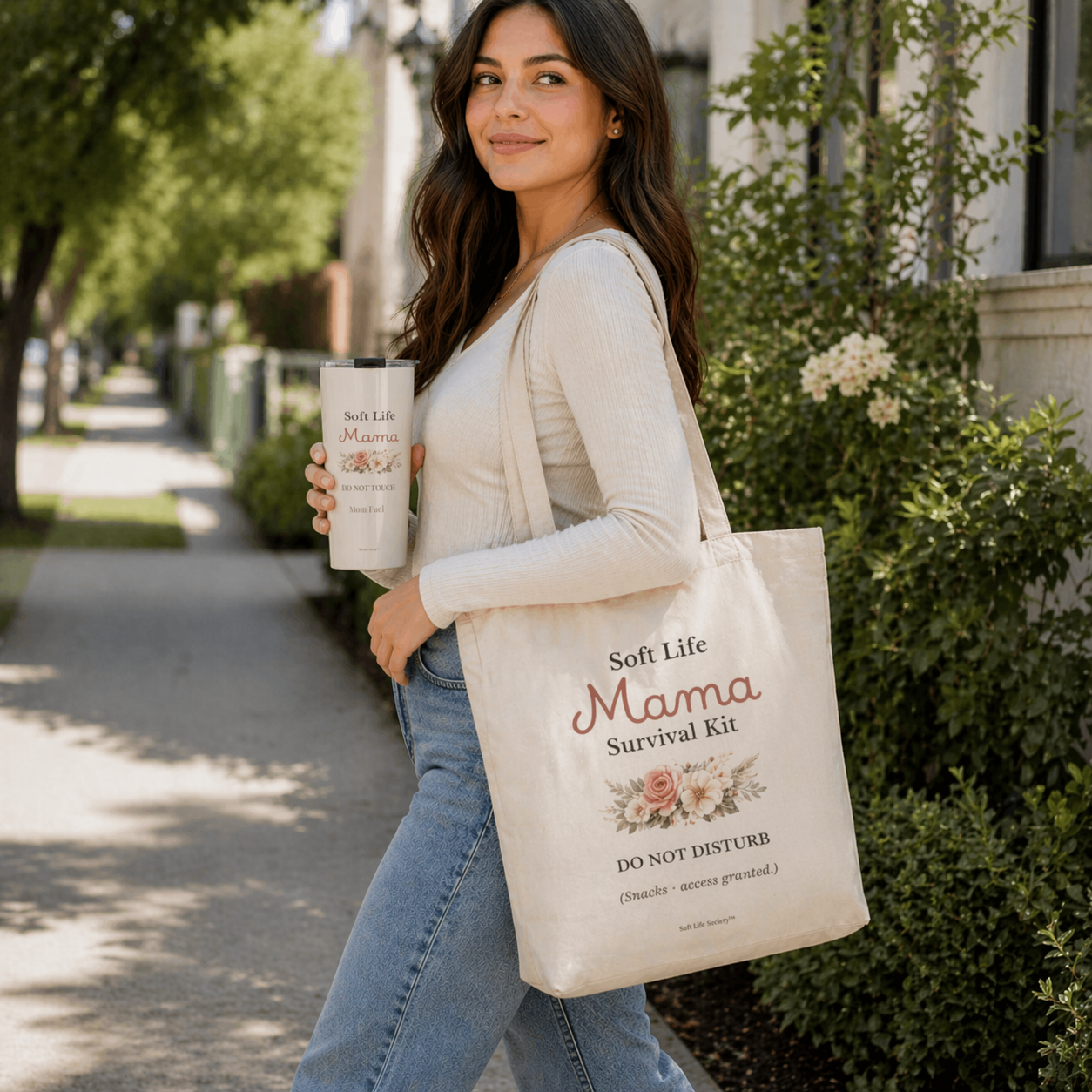Woman holding a tote bag and a mug with 'Soft Life Mama' branding outdoors.