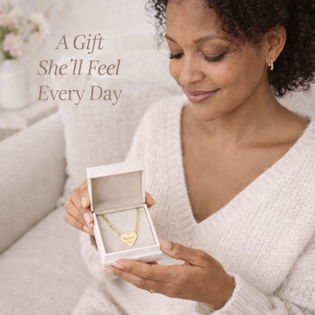 Woman holding a box with a gold heart 'Soft Life Mama' paperclip necklace, sitting on a couch with text 'A Gift She'll Feel Every Day'.