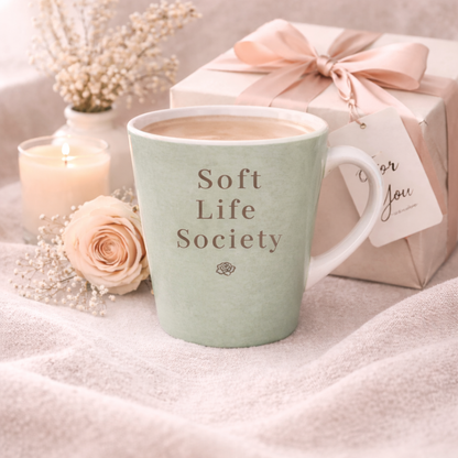 Green mug with 'Soft Life Society' text, gift box, candle, and flowers on a soft surface
