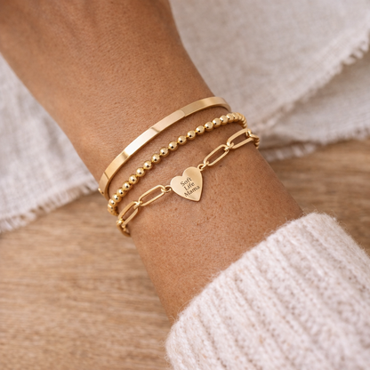 Gold bracelets on a wrist with a heart-shaped engraved pendant reading 'Soft Life Mama', wearing a white sweater.